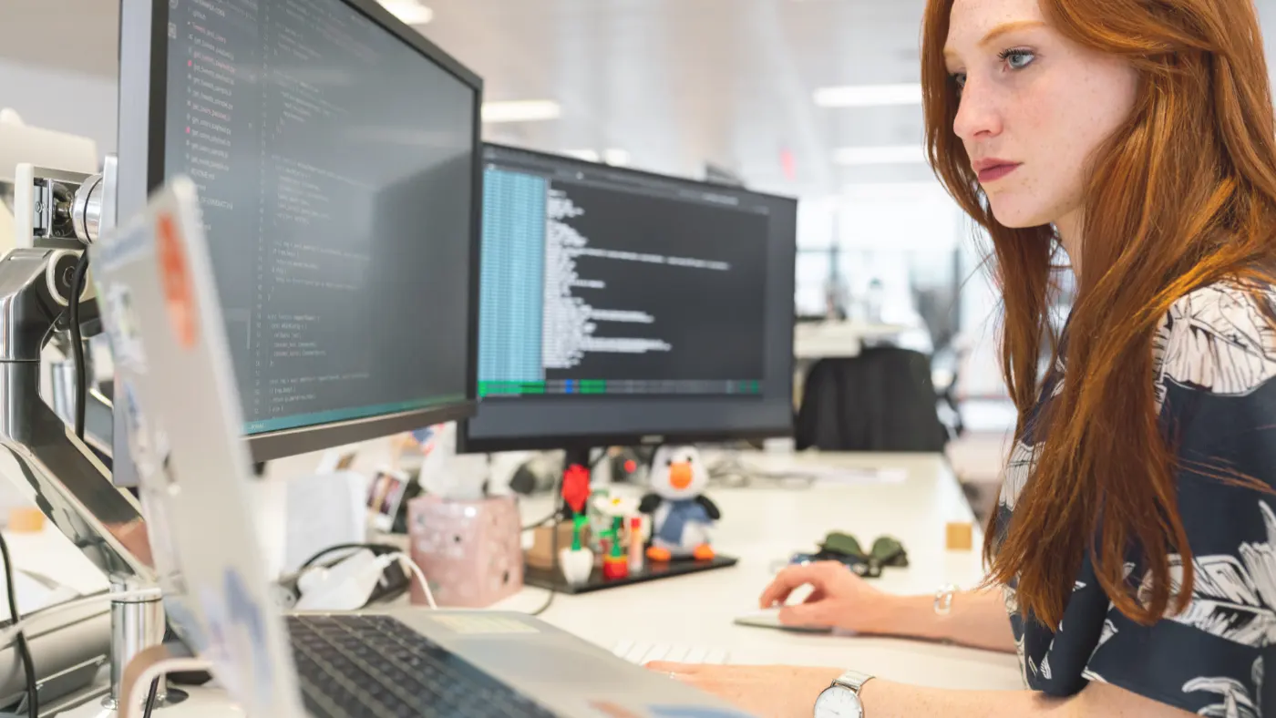 Rödhårig kvinna som sitter vid tre datorskärmar och utvecklar webbplatser. Bild från Pexels-kontot ThisIsEngineering.